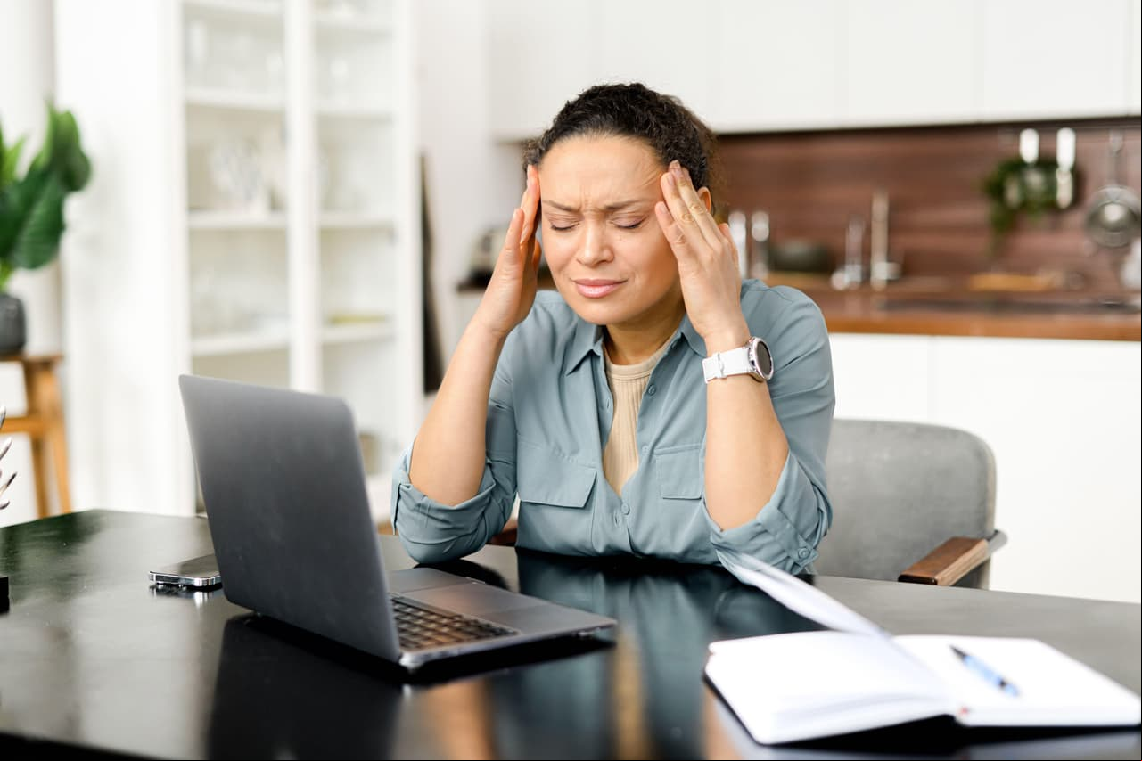 Frustrated young woman using laptop computer suffering from headache, sitting in home office and holding head with hands, feels sick and unwell, burnout and overworked